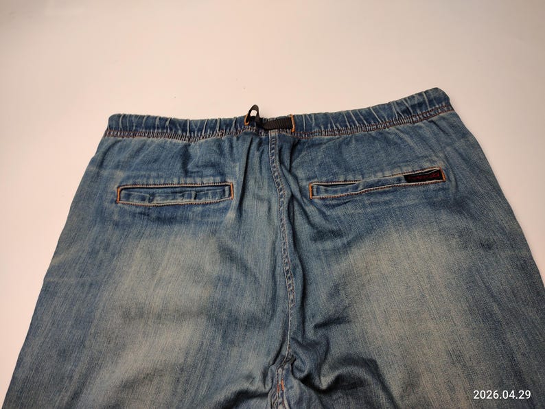 May include: Faded blue denim joggers with an elasticated waistband and drawstring. The back features two pockets with orange stitching and a small, rectangular logo patch. The denim has a worn appearance.