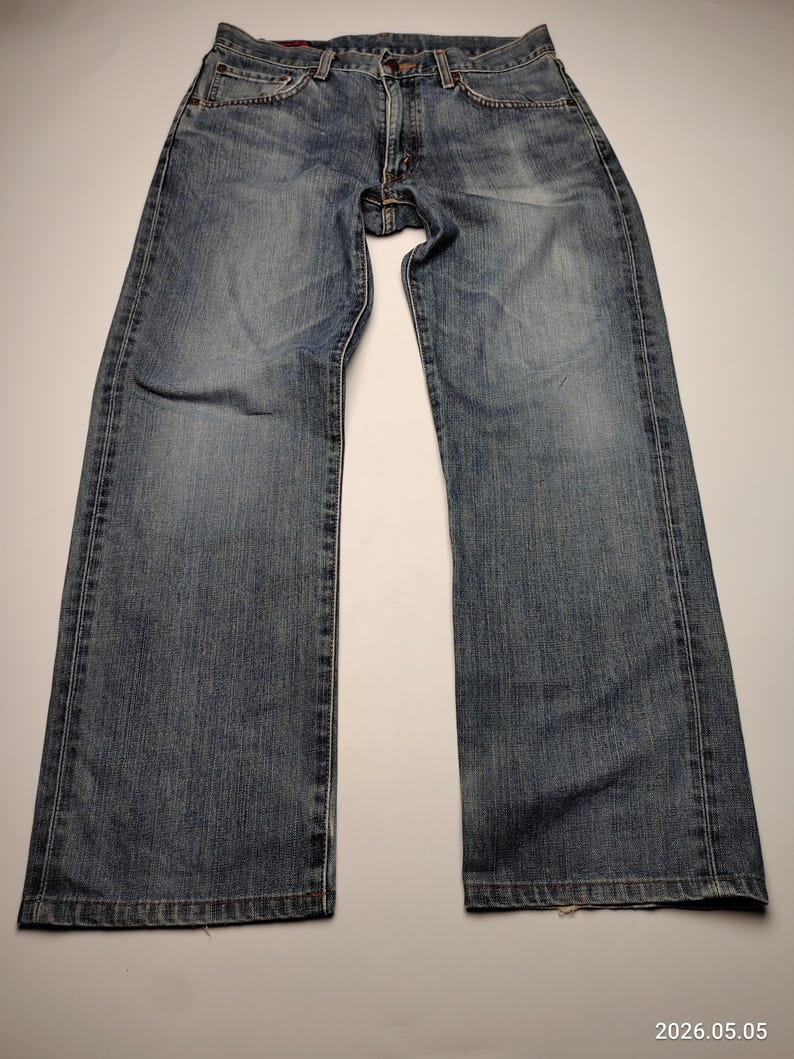 May include: Blue denim jeans with a classic five-pocket design. The jeans have a faded wash and a straight leg cut. The image shows the jeans laid flat on a white surface, showcasing the front and back details.
