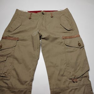 May include: Khaki-colored cargo pants with multiple pockets and brown accents. The pants have a button and zipper closure, and feature a relaxed fit. The image shows the pants laid flat on a white surface.
