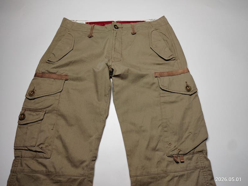 May include: Khaki-colored cargo pants with multiple pockets and brown accents. The pants have a button and zipper closure, and feature a relaxed fit. The image shows the pants laid flat on a white surface.