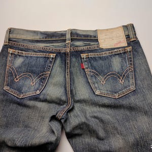 May include: Close-up of the back of blue denim jeans. The jeans have two back pockets with decorative stitching, a red tag, and a leather patch. The denim has a faded, worn appearance.