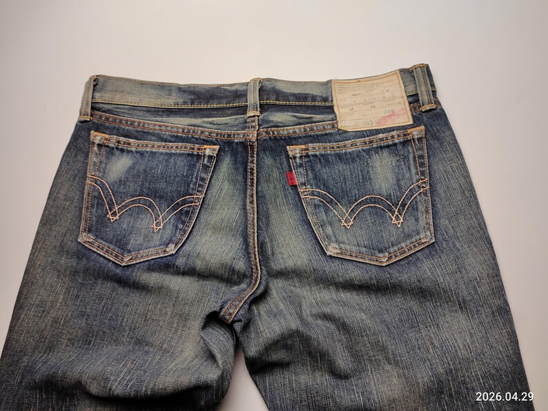 May include: Close-up of the back of blue denim jeans. The jeans have two back pockets with decorative stitching, a red tag, and a leather patch. The denim has a faded, worn appearance.