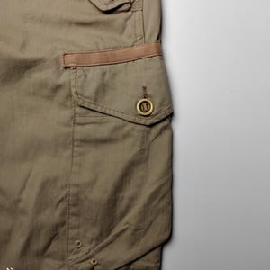 May include: Close-up of olive green cargo pants with a brown accent on the pocket. The pants have a classic design with a button closure and a small pocket. The fabric appears to be a durable cotton blend.