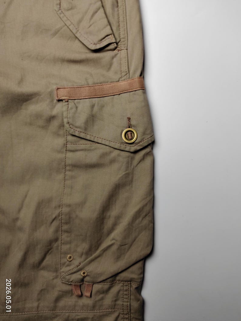 May include: Close-up of olive green cargo pants with a brown accent on the pocket. The pants have a classic design with a button closure and a small pocket. The fabric appears to be a durable cotton blend.