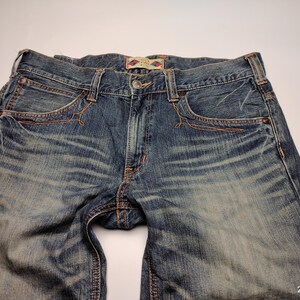 May include: Close-up of blue denim jeans with a faded wash. The jeans have classic five-pocket styling, a button closure, and copper-colored stitching. The fabric shows wear and tear, adding to the vintage aesthetic. The jeans are laid flat.