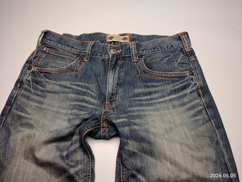May include: Close-up of blue denim jeans with a faded wash. The jeans have classic five-pocket styling, a button closure, and copper-colored stitching. The fabric shows wear and tear, adding to the vintage aesthetic. The jeans are laid flat.