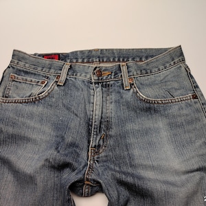 May include: Close-up of blue denim jeans. The jeans have a classic five-pocket design, a button and zipper closure, and copper-colored rivets. The denim has a faded wash, with lighter areas on the thighs and knees.