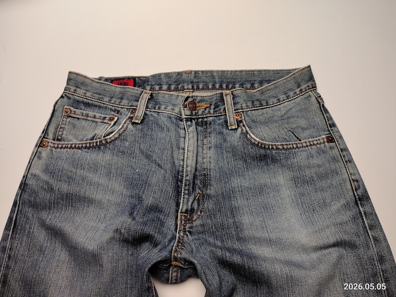 May include: Close-up of blue denim jeans. The jeans have a classic five-pocket design, a button and zipper closure, and copper-colored rivets. The denim has a faded wash, with lighter areas on the thighs and knees.