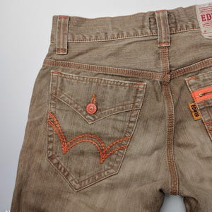 May include: Close-up of brown denim jeans with orange stitching details. The back pocket features a distinctive orange embroidered design. The jeans have a button closure and a brand label. The overall style is casual.
