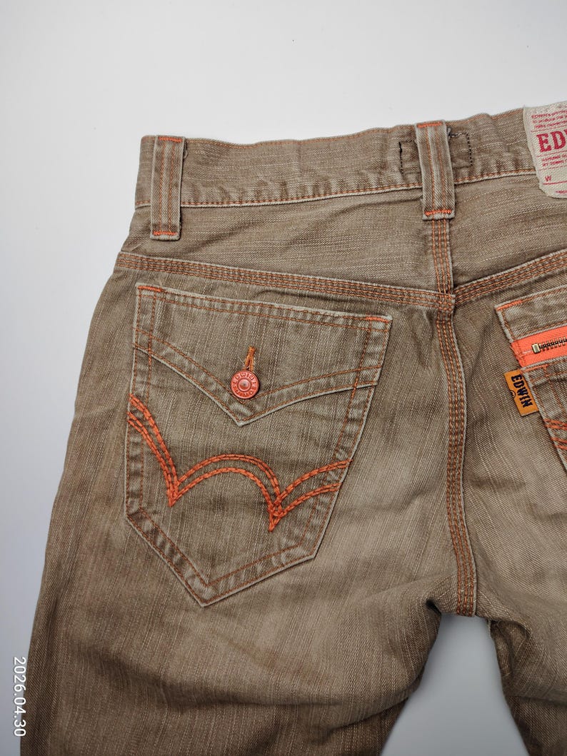 May include: Close-up of brown denim jeans with orange stitching details. The back pocket features a distinctive orange embroidered design. The jeans have a button closure and a brand label. The overall style is casual.