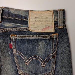 May include: Close-up of blue denim jeans with a back pocket and a red Edwin label. The jeans have a faded wash and visible stitching. A rectangular tag with text is attached to the waistband.