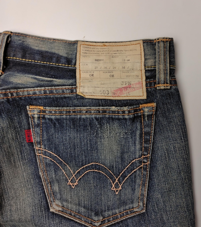 May include: Close-up of blue denim jeans with a back pocket and a red Edwin label. The jeans have a faded wash and visible stitching. A rectangular tag with text is attached to the waistband.