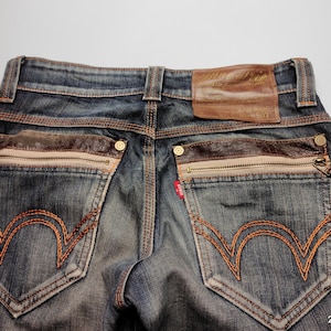 Puede incluir: Primer plano de la parte trasera de unos vaqueros azules con detalles en cuero marr&oacute;n. Incluye bolsillos con cremallera, costuras decorativas y un parche de cuero. Los vaqueros tienen un lavado descolorido y un dise&ntilde;o cl&aacute;sico de cinco bolsillos.