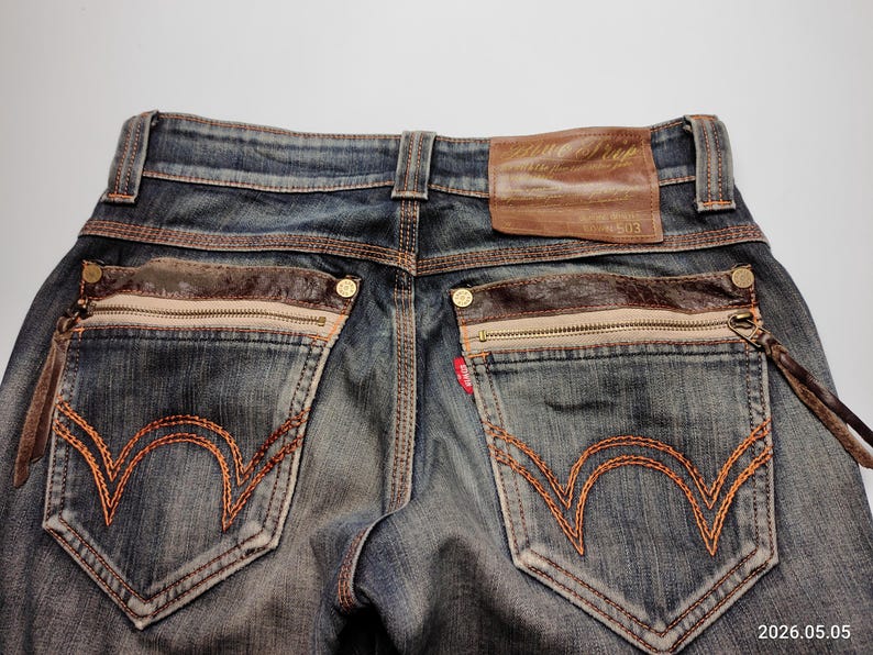 Puede incluir: Primer plano de la parte trasera de unos vaqueros azules con detalles en cuero marr&oacute;n. Incluye bolsillos con cremallera, costuras decorativas y un parche de cuero. Los vaqueros tienen un lavado descolorido y un dise&ntilde;o cl&aacute;sico de cinco bolsillos.