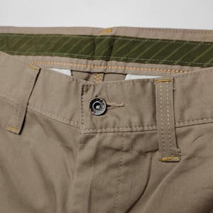 May include: Close-up of khaki-colored pants. The pants feature a button closure, belt loops, and a green and khaki waistband. The stitching is visible in a contrasting yellow thread. The fabric appears to be a durable cotton blend.