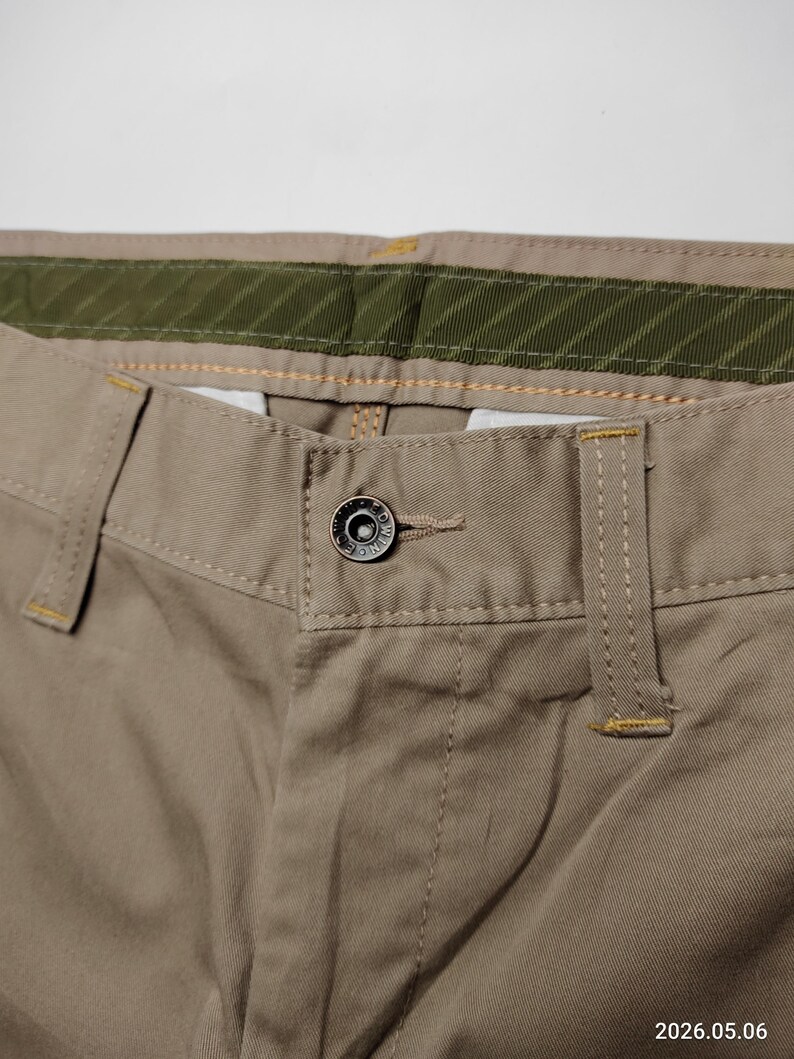 May include: Close-up of khaki-colored pants. The pants feature a button closure, belt loops, and a green and khaki waistband. The stitching is visible in a contrasting yellow thread. The fabric appears to be a durable cotton blend.