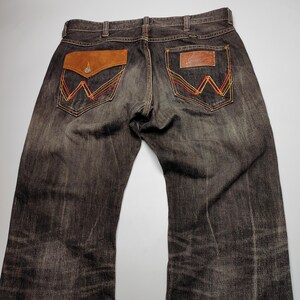 May include: Dark brown denim jeans with a faded wash. The back pockets feature a signature "W" design with orange, red, and yellow stitching. One pocket has a brown suede flap with a button. A leather patch is on the other pocket.