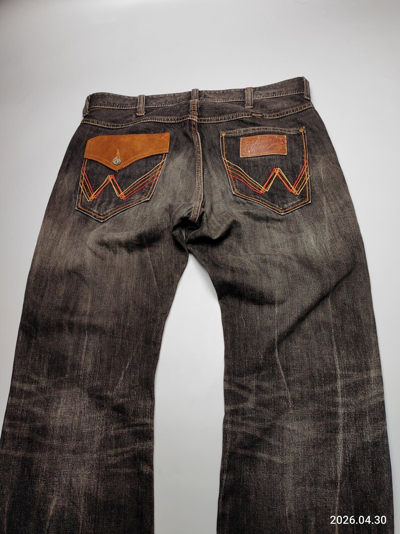 May include: Dark brown denim jeans with a faded wash. The back pockets feature a signature "W" design with orange, red, and yellow stitching. One pocket has a brown suede flap with a button. A leather patch is on the other pocket.