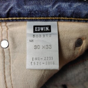 May include: Close-up of a denim jeans label with the brand name "EDWIN." The label displays "553 RV2" and size "30 x 33." Additional codes "EME-2299" and "E520-2006" are also visible. The jeans are blue.