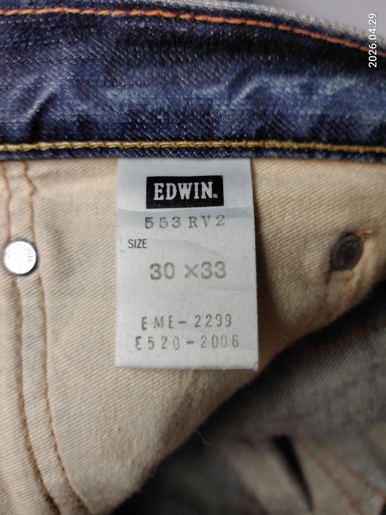 May include: Close-up of a denim jeans label with the brand name "EDWIN." The label displays "553 RV2" and size "30 x 33." Additional codes "EME-2299" and "E520-2006" are also visible. The jeans are blue.