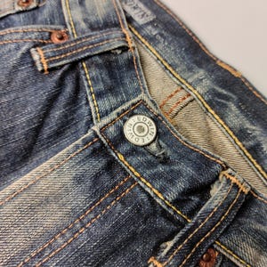 May include: Close-up of blue denim jeans with visible stitching in a contrasting orange-yellow thread. The jeans feature a metal button with the brand name "EDWIN" and copper rivets. The fabric shows signs of wear and fading.