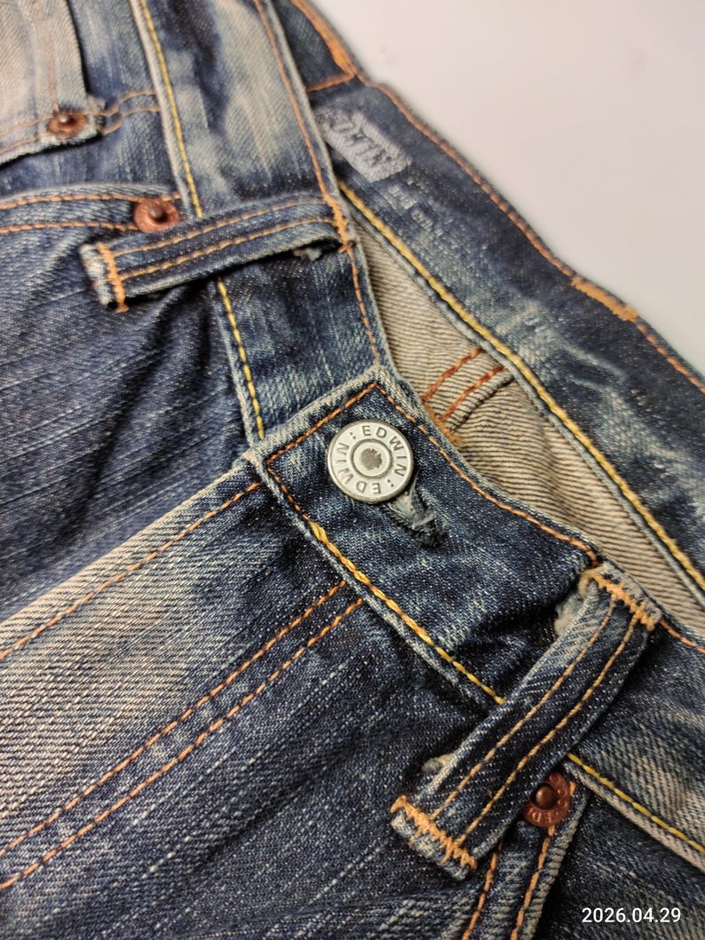 May include: Close-up of blue denim jeans with visible stitching in a contrasting orange-yellow thread. The jeans feature a metal button with the brand name "EDWIN" and copper rivets. The fabric shows signs of wear and fading.