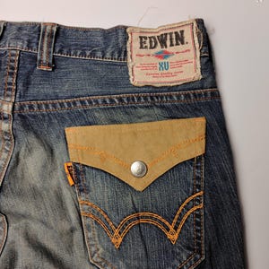 May include: Close-up of blue denim jeans with a light brown suede pocket and orange stitching. The back pocket has a unique design. The Edwin brand label is visible at the top.