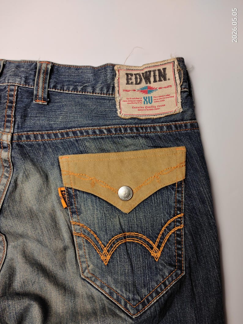 May include: Close-up of blue denim jeans with a light brown suede pocket and orange stitching. The back pocket has a unique design. The Edwin brand label is visible at the top.