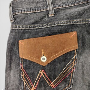May include: Close-up of the back pocket of a pair of gray denim jeans. The pocket features a brown suede flap with a button closure. The pocket is decorated with orange, pink, and white stitching in a geometric pattern. The jeans have a classic five-pocket design.