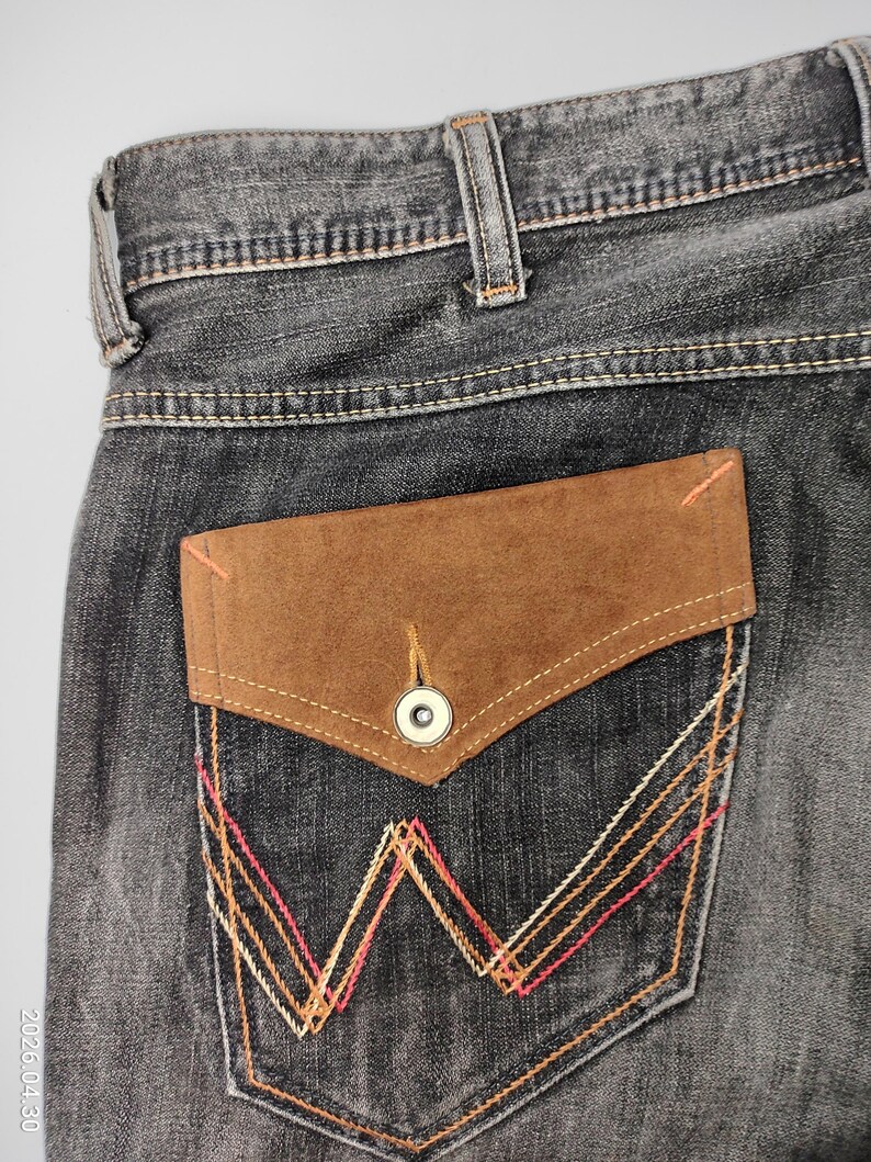 May include: Close-up of the back pocket of a pair of gray denim jeans. The pocket features a brown suede flap with a button closure. The pocket is decorated with orange, pink, and white stitching in a geometric pattern. The jeans have a classic five-pocket design.