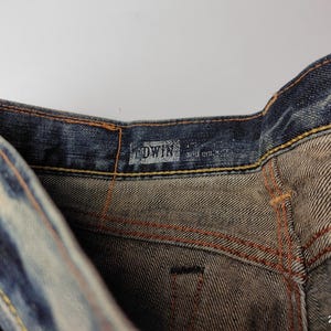 May include: Close-up of denim jeans with visible stitching and the brand name "EDWIN" on the waistband. The jeans have a faded blue wash with lighter areas, and the stitching is in a contrasting orange color.