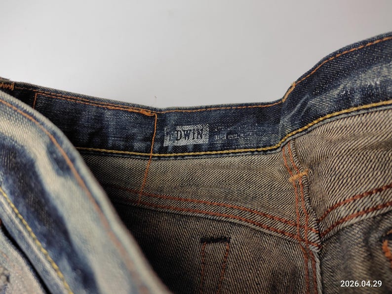 May include: Close-up of denim jeans with visible stitching and the brand name "EDWIN" on the waistband. The jeans have a faded blue wash with lighter areas, and the stitching is in a contrasting orange color.