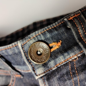 Puede incluir: Primer plano de unos vaqueros azules. Los vaqueros presentan un bot&oacute;n circular decorativo con un dise&ntilde;o floral. Las costuras y el ojal est&aacute;n resaltados con pespuntes naranjas. La tela tiene un aspecto desgastado y descolorido.