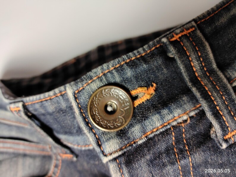Puede incluir: Primer plano de unos vaqueros azules. Los vaqueros presentan un bot&oacute;n circular decorativo con un dise&ntilde;o floral. Las costuras y el ojal est&aacute;n resaltados con pespuntes naranjas. La tela tiene un aspecto desgastado y descolorido.