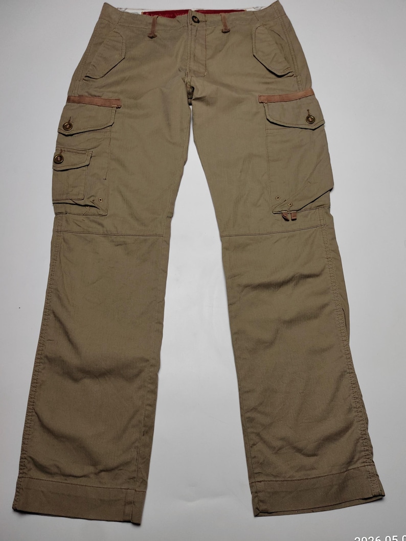 May include: Khaki-colored cargo pants with multiple pockets and button closures. The pants have a relaxed fit and feature reinforced details at the pockets. The design includes a button and zipper fly, and a brown accent on the pockets.
