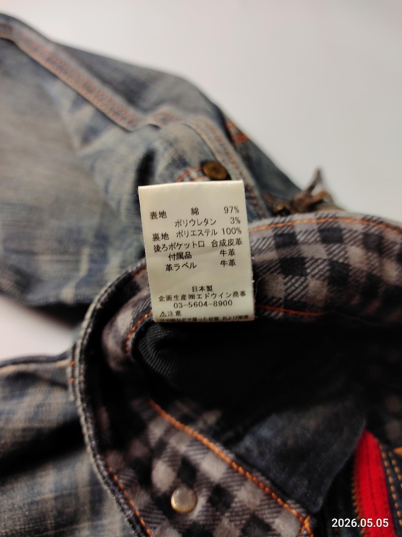Puede incluir: Primer plano de una chaqueta vaquera con forro de cuadros visible. La chaqueta tiene costuras visibles y una etiqueta con texto japon&eacute;s. La etiqueta enumera los materiales: algod&oacute;n, poliuretano y poli&eacute;ster.