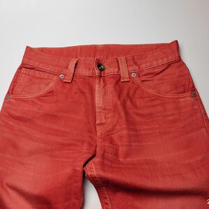 May include: Close-up of red denim jeans. The jeans have a classic five-pocket design with a button and zipper closure. The fabric has a slightly faded appearance, and the stitching is visible in a contrasting color.