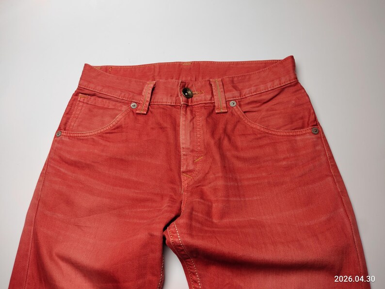 May include: Close-up of red denim jeans. The jeans have a classic five-pocket design with a button and zipper closure. The fabric has a slightly faded appearance, and the stitching is visible in a contrasting color.
