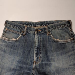 Peut inclure: Gros plan sur un jean en denim bleu. Le jean pr&eacute;sente une conception classique &agrave; cinq poches avec une fermeture boutonn&eacute;e et zipp&eacute;e. Le denim a un aspect d&eacute;lav&eacute; et us&eacute;, avec une usure visible autour des poches et des bords, lui donnant un aspect vintage.