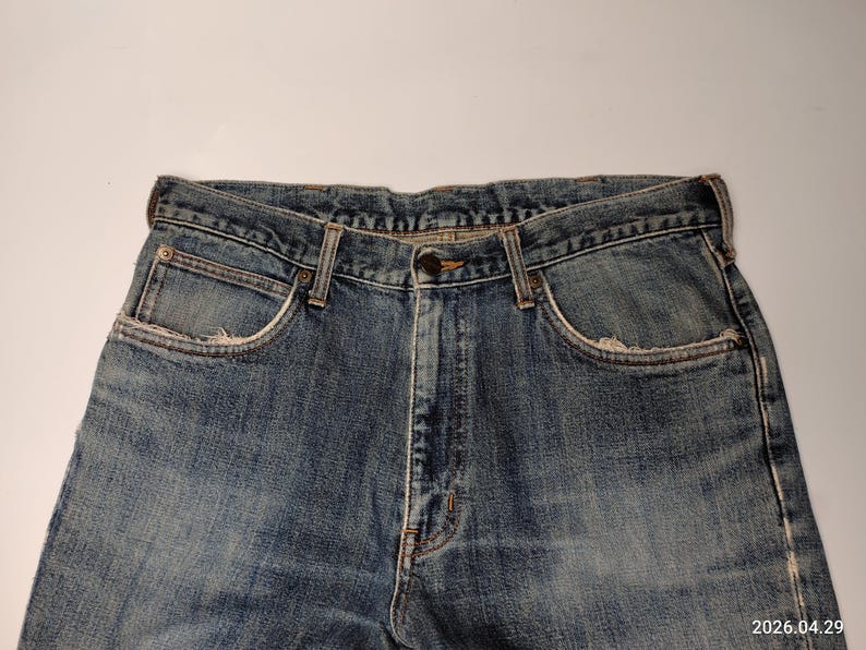 Peut inclure: Gros plan sur un jean en denim bleu. Le jean pr&eacute;sente une conception classique &agrave; cinq poches avec une fermeture boutonn&eacute;e et zipp&eacute;e. Le denim a un aspect d&eacute;lav&eacute; et us&eacute;, avec une usure visible autour des poches et des bords, lui donnant un aspect vintage.