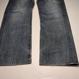 May include: A close-up shot of the lower portion of a pair of blue denim jeans. The jeans are faded and show signs of wear, with visible seams and a classic five-pocket design. The denim has a medium wash.