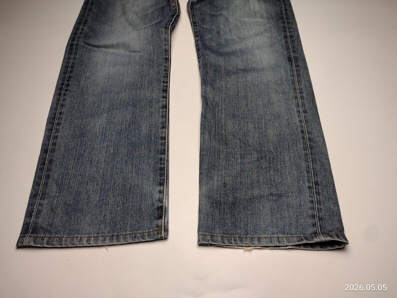 May include: A close-up shot of the lower portion of a pair of blue denim jeans. The jeans are faded and show signs of wear, with visible seams and a classic five-pocket design. The denim has a medium wash.