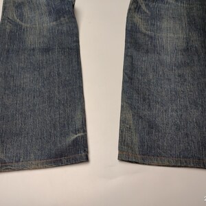 May include: Close-up of the lower portion of a pair of blue denim jeans. The jeans have a faded appearance with visible wear and tear, and the hems are visible. The denim has a classic, textured look.