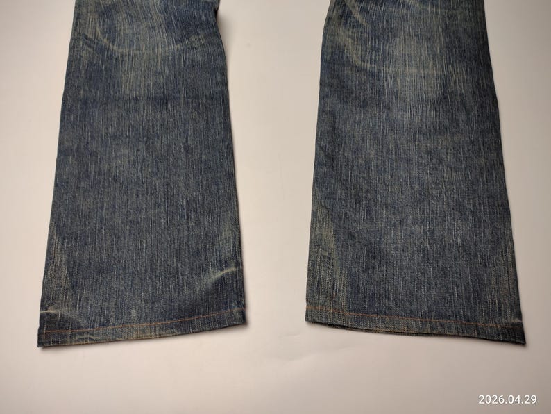 May include: Close-up of the lower portion of a pair of blue denim jeans. The jeans have a faded appearance with visible wear and tear, and the hems are visible. The denim has a classic, textured look.