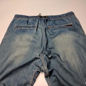 May include: Light blue denim trousers with an elasticated waistband and drawstring. The trousers feature two back pockets and a small logo patch. The denim has a faded, worn appearance.