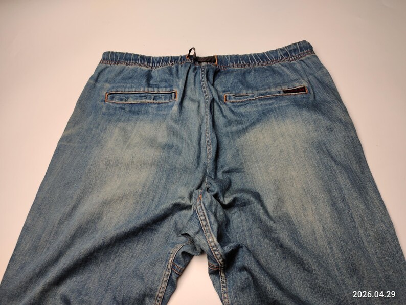 May include: Light blue denim trousers with an elasticated waistband and drawstring. The trousers feature two back pockets and a small logo patch. The denim has a faded, worn appearance.