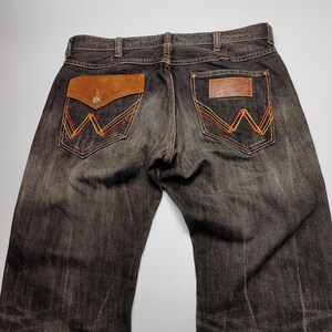 May include: Dark wash denim jeans with brown leather pocket accents and decorative stitching. The back pockets feature a classic 'W' design in red, orange, and yellow thread. The jeans have a button closure and belt loops.