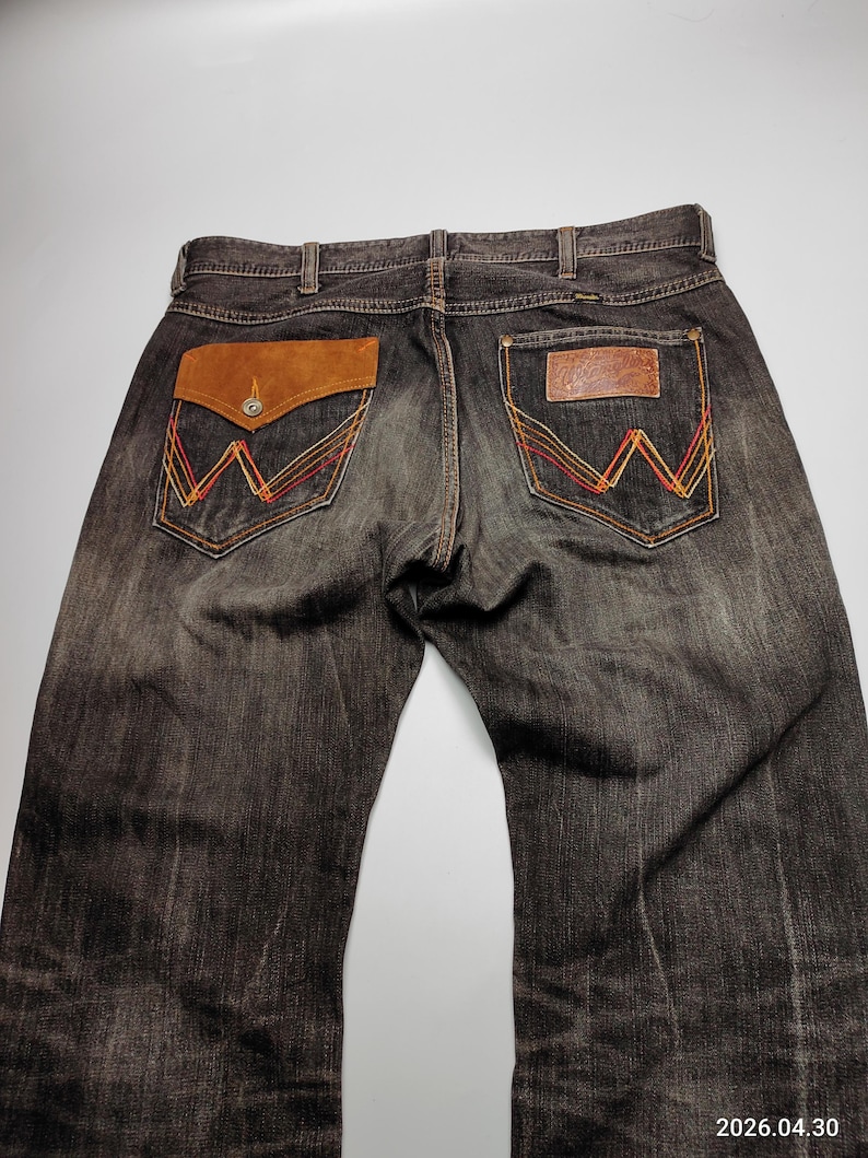 May include: Dark wash denim jeans with brown leather pocket accents and decorative stitching. The back pockets feature a classic 'W' design in red, orange, and yellow thread. The jeans have a button closure and belt loops.