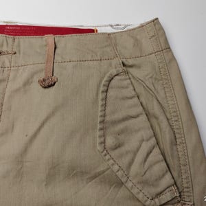 May include: Close-up of tan-colored shorts with a button closure and a small pocket. The shorts have visible stitching and a textured fabric. The shorts are made of a durable material.