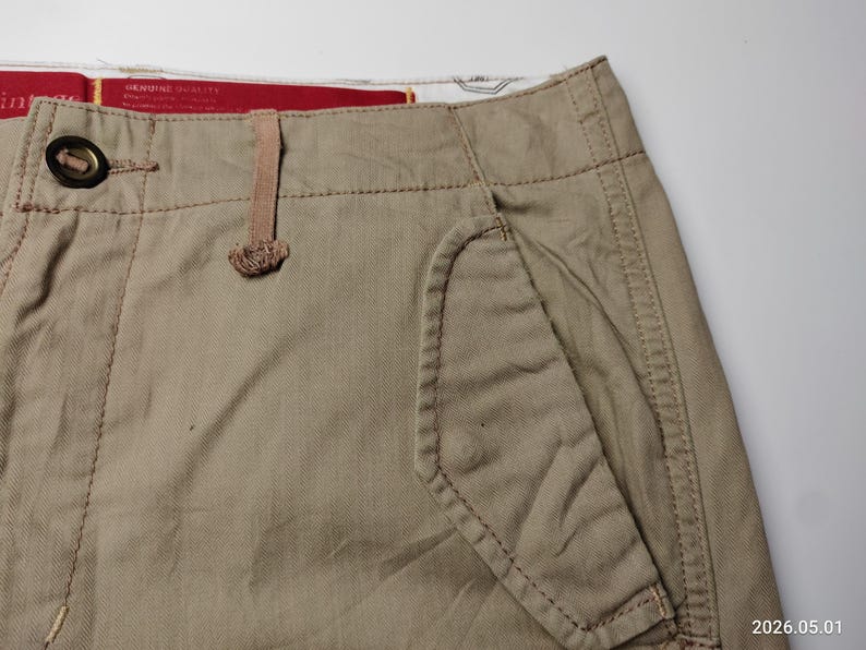 May include: Close-up of tan-colored shorts with a button closure and a small pocket. The shorts have visible stitching and a textured fabric. The shorts are made of a durable material.
